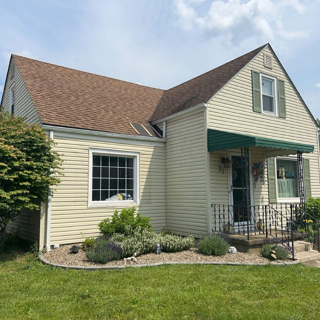 A cozy yellow house featuring a brown roof, green awning, and vibrant garden. The front door is adorned with flower baskets, nestled in a suburban setting.