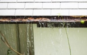 Overflowing dirty gutter filled with leaves and debris spills water onto the exterior green siding of a home.