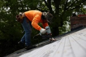 A roofer wearing an orange shirt installs asphalt shingles on a sloped roof using a power nailer. The background shows green trees and a brick chimney.