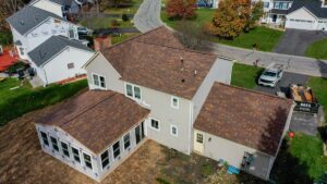 Two-story beige house with a brown shingled roof and an attached garage. Aerial view showing neighboring properties and paved roads.