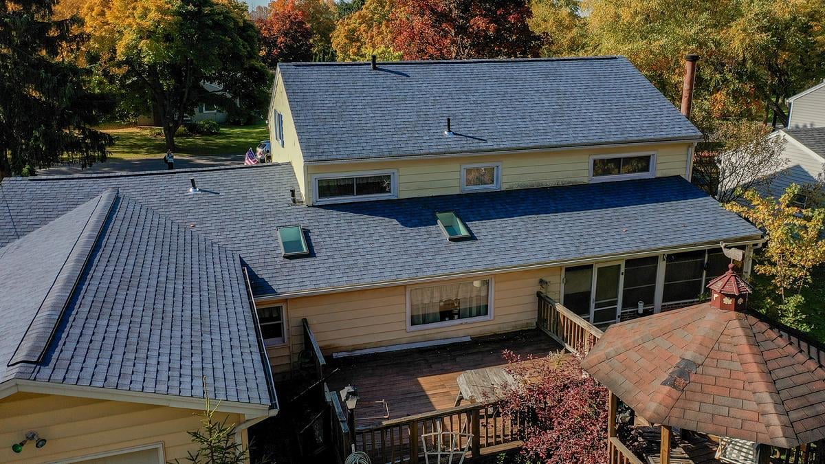 An aerial view of a residential house featuring blue asphalt shingle roof, autumn-colored trees, and a adjacent gazebo structure.