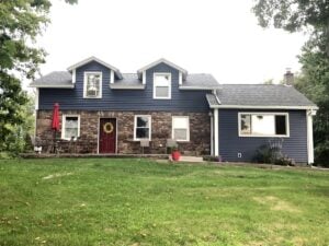 A two-story house with blue siding and brown stone foundation featuring white-trimmed windows and a red front door, set against a green lawn and trees.