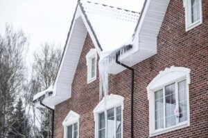 A brick house with snow-covered roof and large icicles hanging from the gutter. Bare winter trees are visible in the background.