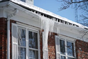 A red brick house with snow-covered gutters and large icicles hanging down against a clear blue sky and bare trees.