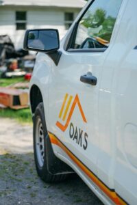 A clean white company truck featuring a prominent orange DAKS logo with a house icon, parked near a residential house and a trailer.