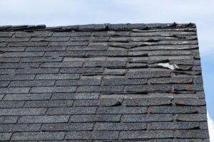 Weathered asphalt shingles on a roof showing signs of lifting, cracking, and wear against a pale sky.