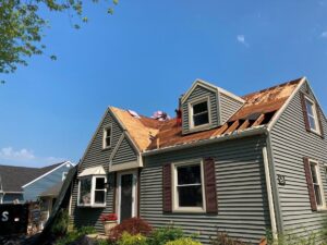 A gray house with a partially stripped roof showing wood underneath under a clear blue sky.