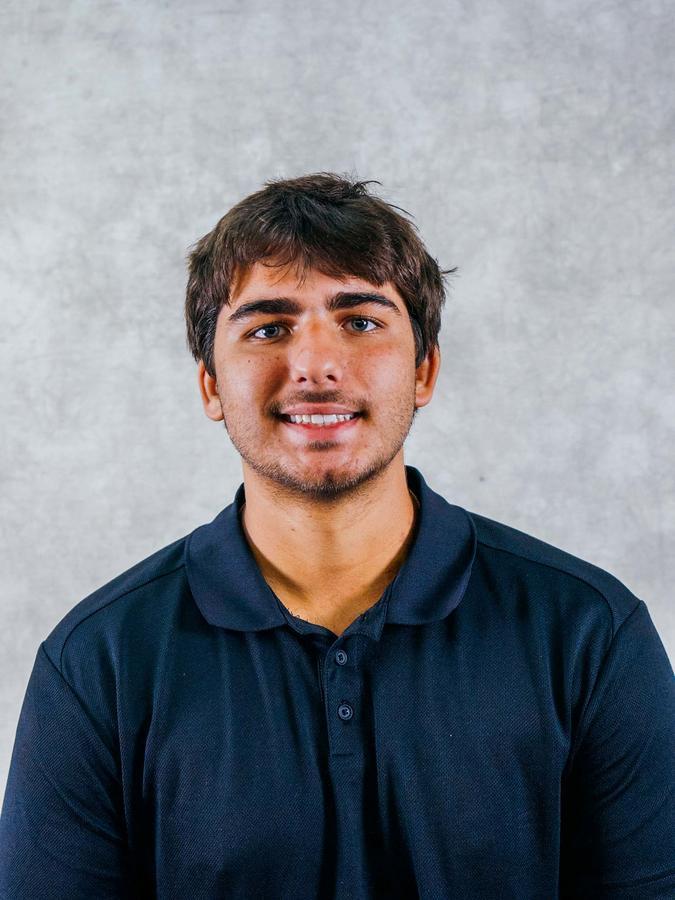 Portrait of a smiling young man with short brown hair and a dark polo shirt. He is standing against a gray mottled background.