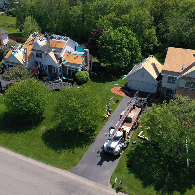 An aerial view of a neighborhood with houses undergoing roofing work. Workers and equipment are visible on the roofs and in the driveways.