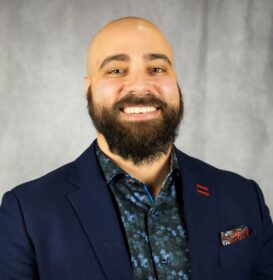 A bald man with a beard wearing a blue suit and smiling. He has a red pocket square.