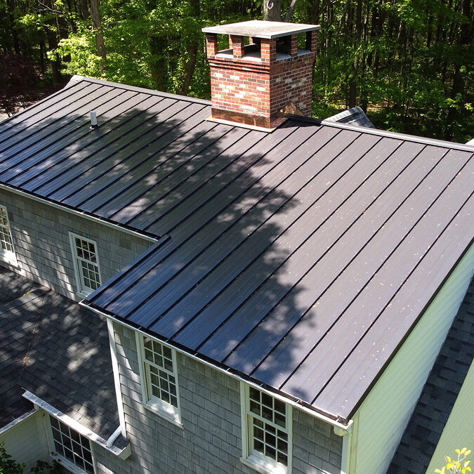 A gray metal roof on a house with a brick chimney. The roof is surrounded by trees.