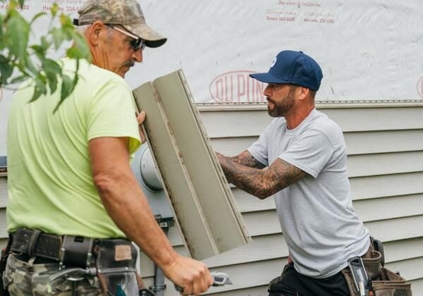 Two workers in caps and shorts holding siding panel. They are working on house exterior.