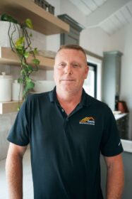 A man wearing a black polo shirt with a company logo on the left side of his chest. He appears to be standing in a modern kitchen or office.