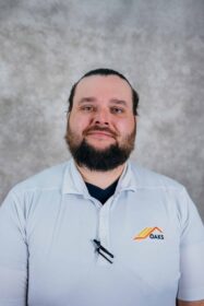 A man with a beard wearing a white polo shirt. He is standing in front of a gray background.