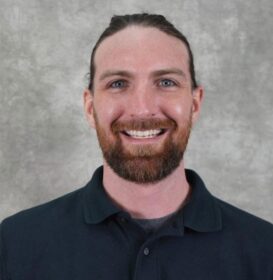 A man with a beard and a smile. He is wearing a dark shirt.