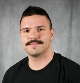 A man with a moustache wearing a black shirt. He has short dark hair.