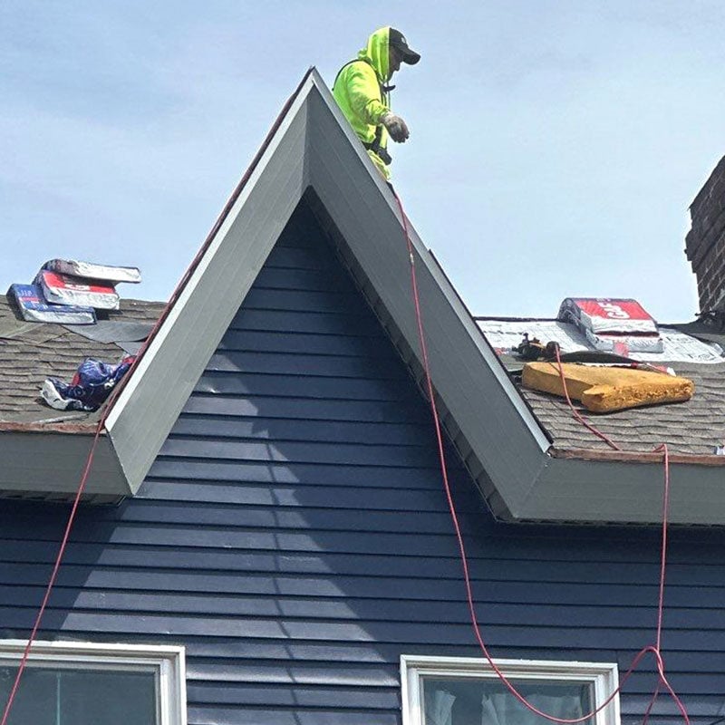 Roofing Work in Progress on Blue House A person in a yellow vest working on a roof. Roofing materials are scattered around.