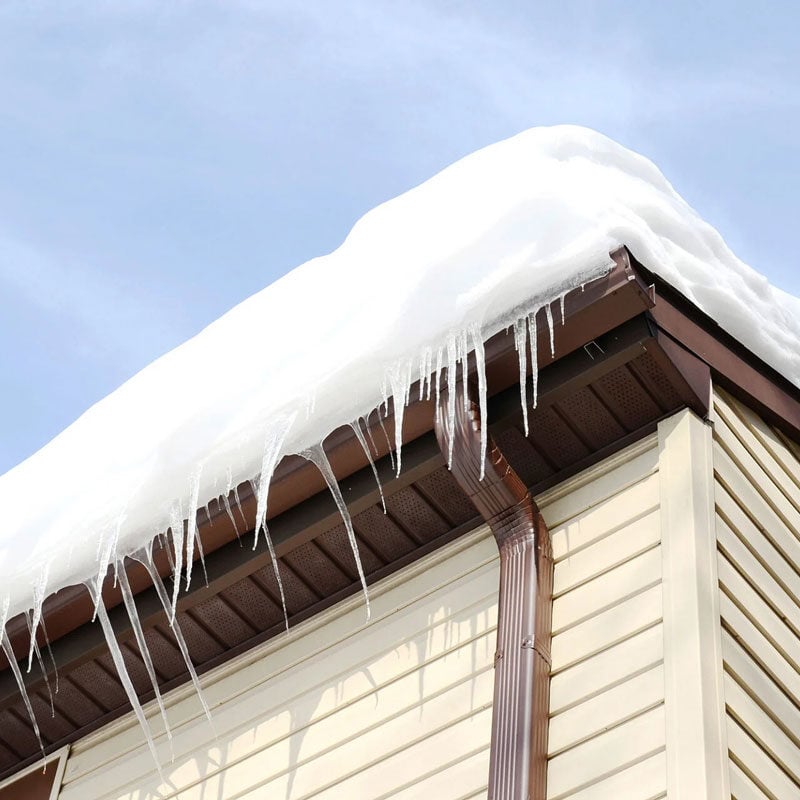 Icicles hanging from a snowy roof edge. Roof and gutter clogged with snow.
