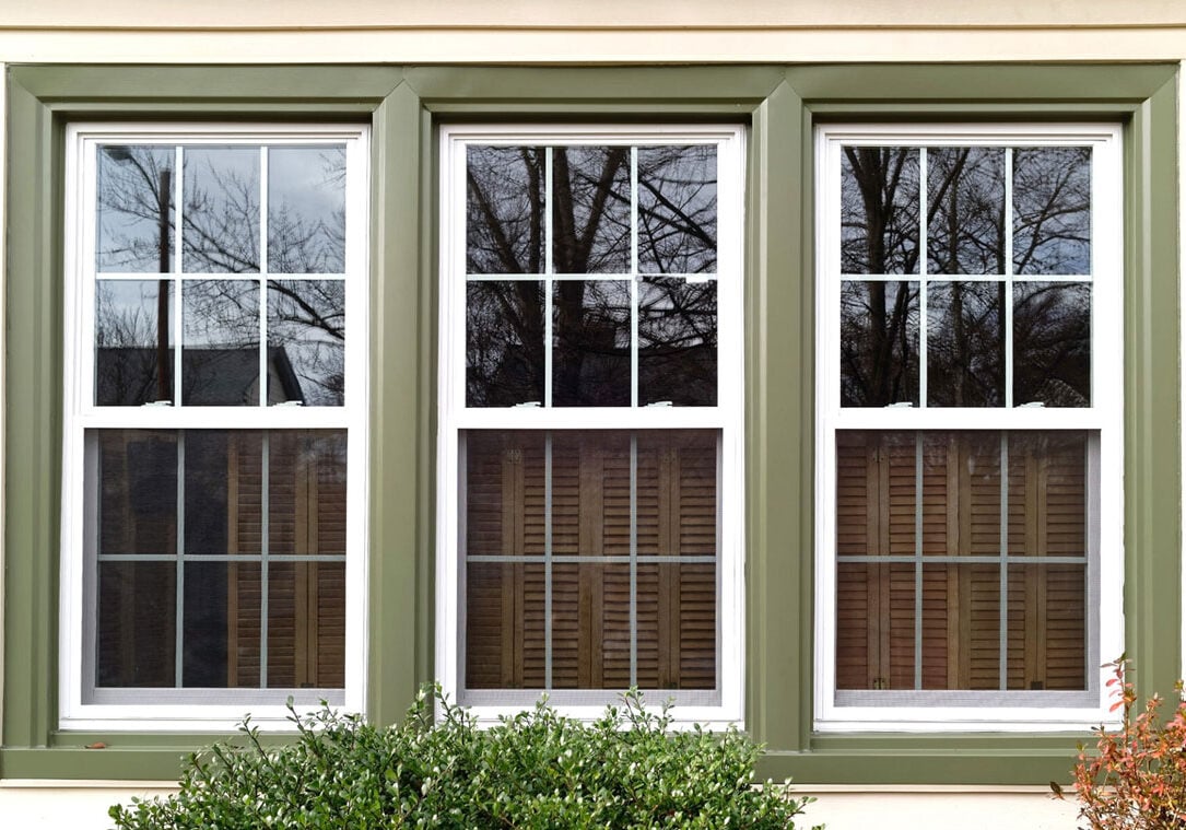 Three Windows with Green Trim and White Frames A set of three white framed windows with green trim reflecting trees and sky. Brown blinds are partially closed.